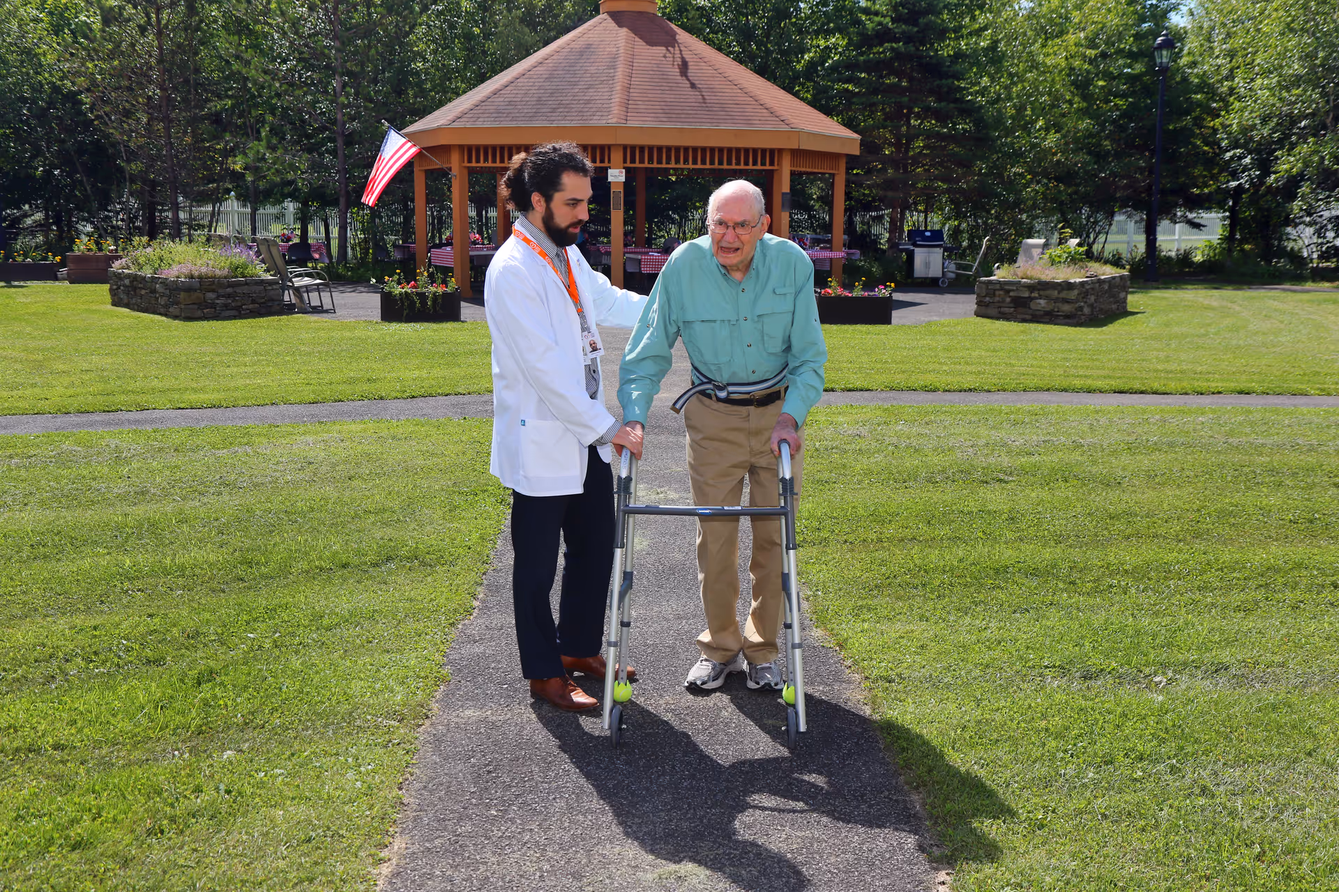 An elderly man using a walker is being assisted by a healthcare professional wearing a white coat outdoors on a paved path with green grass and trees around. In the background, there is a wooden gazebo with an American flag and some chairs and tables.