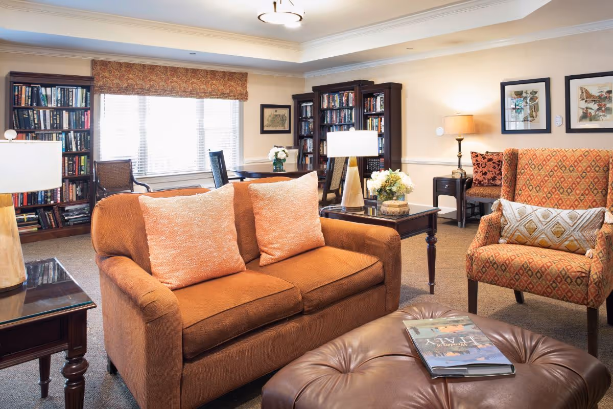 A cozy living room area with an orange loveseat with two matching pillows, a patterned armchair with a decorative pillow, a large leather ottoman with a book on it, two wooden side tables with lamps, bookshelves filled with books, a window with blinds and a valance, and framed artwork on the walls.
