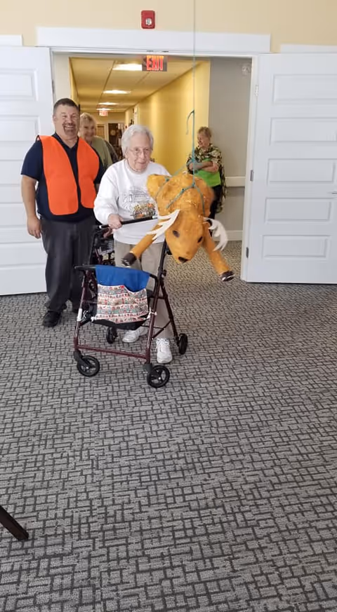An elderly woman using a walker is holding a stuffed moose toy suspended by a string in a hallway. Behind her, a man wearing an orange safety vest and two other elderly women are standing and smiling. The setting appears to be inside a senior living facility with carpeted floors and white double doors.