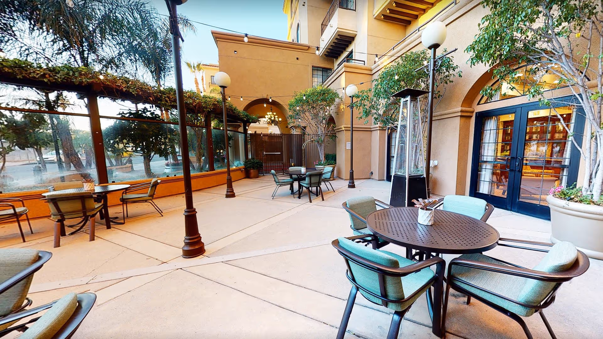 Outdoor patio area at Atria Tarzana with round tables and cushioned chairs arranged for seating. The space is surrounded by a building with arched doorways and large windows, potted plants, tall street lamps, and a patio heater. Trees and greenery are visible along the perimeter.