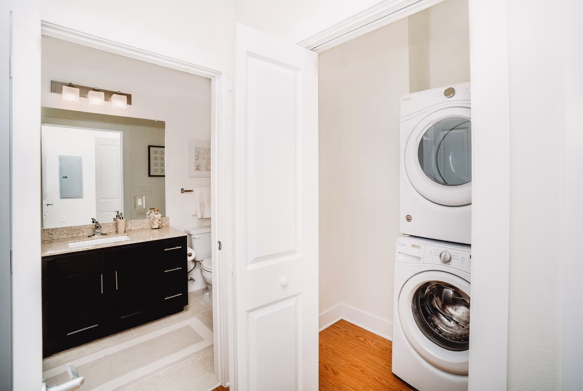 View of a bathroom with a dark wood vanity, granite countertop, sink, mirror, and light fixture above. Next to the bathroom is a small laundry area with a stacked white washer and dryer. The floor in the laundry area is wood, and the bathroom floor is tiled.