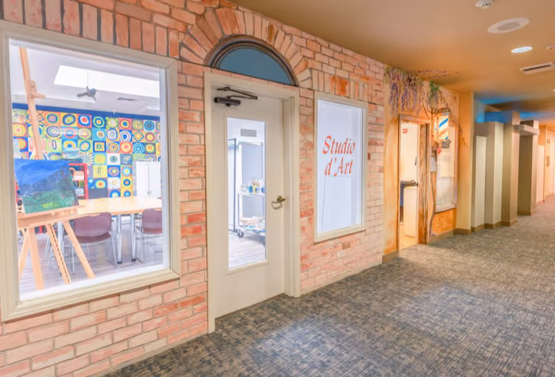 Interior hallway of Cherrywood Village a Generations Community showing the entrance to an art studio with large windows. Inside the studio, there are tables, chairs, an easel with a painting, and a colorful wall with circular patterns. The hallway has carpeted flooring and brick-style walls with an arched doorway.