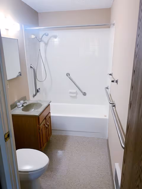 Small bathroom with a white bathtub and shower combination, white tiled wall surround, a silver grab bar on the wall, a wooden vanity with a sink and faucet, a mirror above the sink, a toilet, and beige walls with a towel bar on the right side.