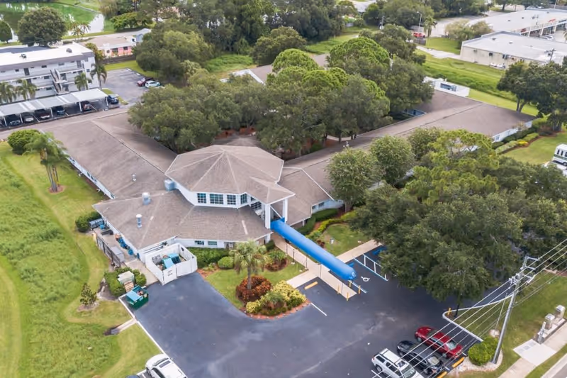 Aerial view of The Pointe senior living facility showing a large single-story building with a central covered entrance, surrounded by trees and greenery. There is a parking lot with several cars and a blue canopy over the main entrance walkway.