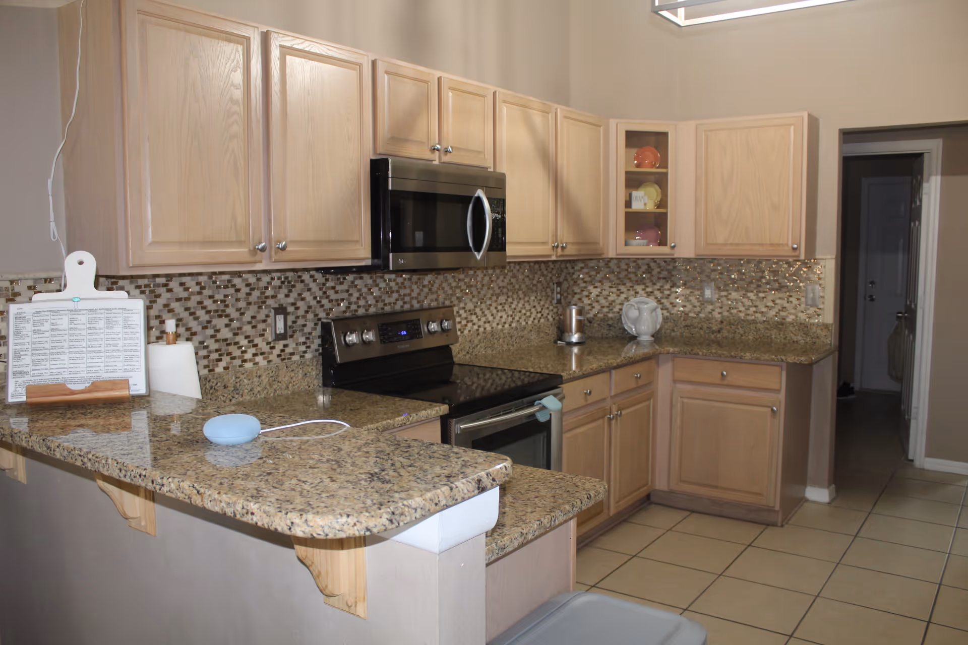 A kitchen with light wood cabinets, granite countertops, and a mosaic tile backsplash. The kitchen features a stainless steel microwave and stove, a paper towel holder, a small blue smart speaker, and some decorative dishes in a glass-front cabinet. The floor is tiled, and there is a doorway leading to another room.