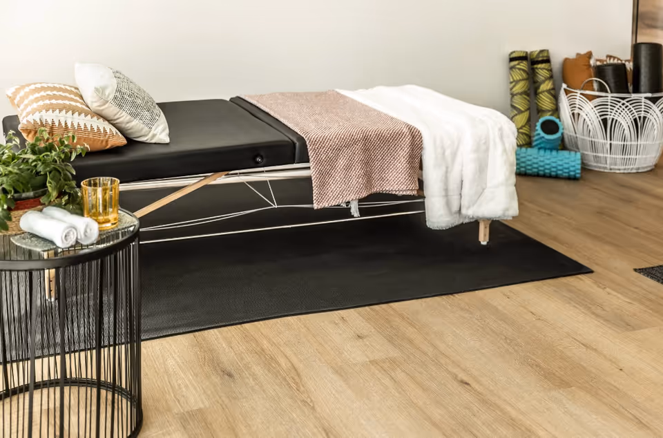 A modern daybed with pillows and throws next to a small round side table and exercise mats on a wood floor.