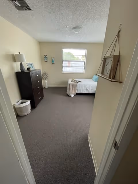 View into a simple bedroom with beige walls and carpeted floor. The room contains a single bed with white bedding and a light blue pillow, positioned under a window with blinds. To the left, there is a dark wooden chest of drawers with a lamp and decorative items on top. A white basket is placed on the floor next to the chest. A small wall hanging is visible on the right wall near the entrance.