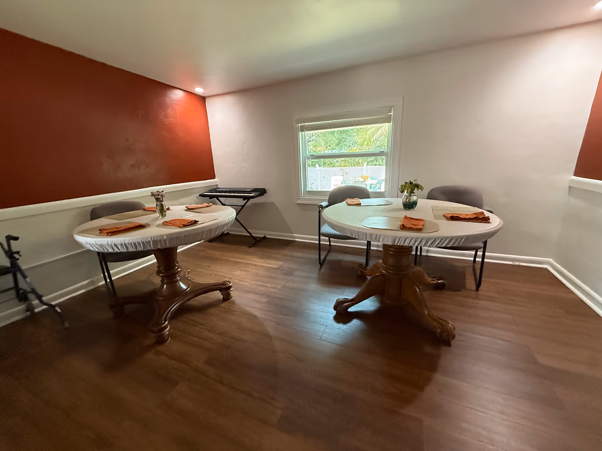 A small dining area with two round wooden tables covered with white tablecloths, each set with placemats and orange napkins. There are chairs around the tables, a window showing greenery outside, and a keyboard on a stand against the wall. The walls are painted white with a red accent wall, and the floor is wooden.