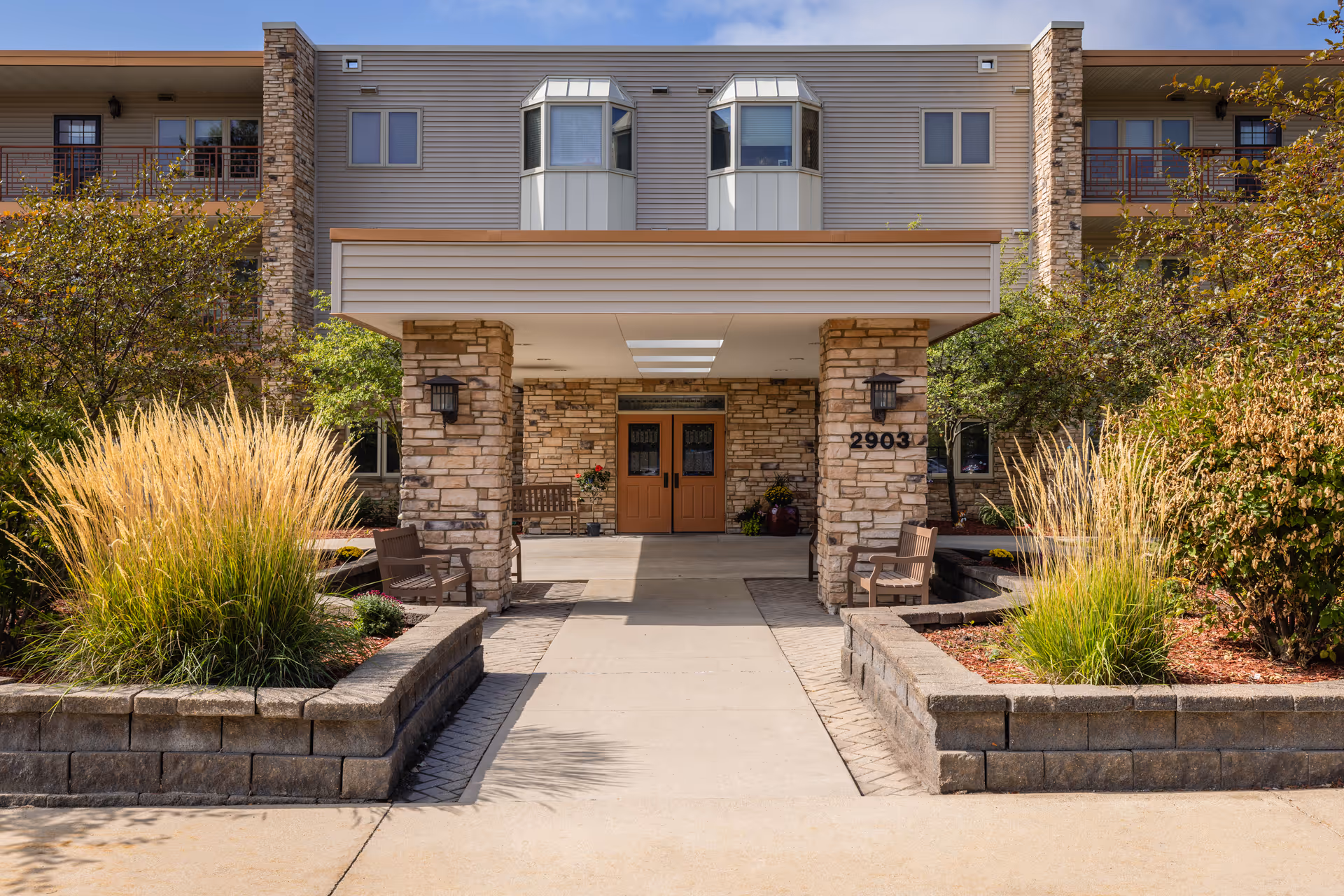 Entrance of a senior living facility with a covered walkway supported by stone pillars, flanked by landscaped areas with ornamental grasses and shrubs. There are benches on either side near the entrance doors, and the building has a beige and stone exterior with balconies on the upper floors.