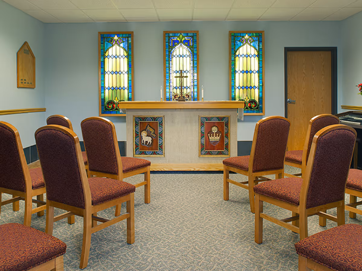 Small chapel with upholstered chairs facing an altar and three stained-glass windows.