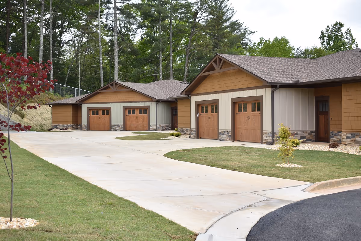 Exterior view of a senior living facility with multiple attached garages featuring wooden doors, surrounded by a well-maintained lawn and trees in the background.