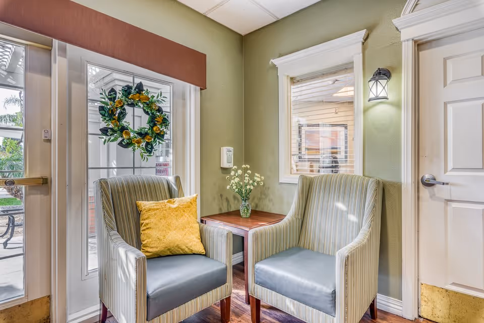 A cozy corner seating area with two striped armchairs, one with a yellow cushion, positioned around a small wooden table with a vase of white flowers. The space has green walls, a window with white blinds, a door with a silver handle, and a glass door decorated with a green and yellow wreath. A wall-mounted light fixture is above the window.