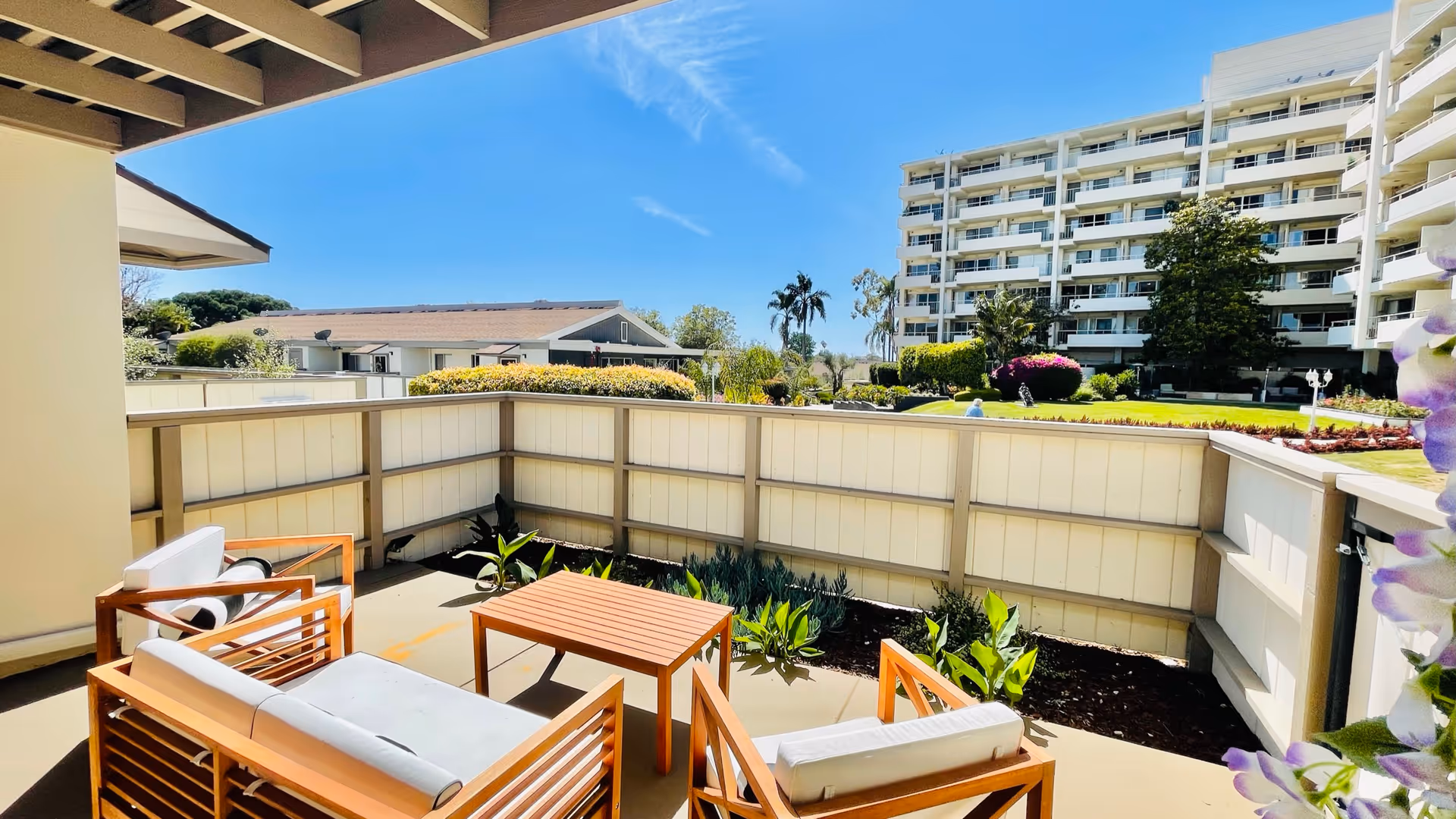 Outdoor patio area with wooden outdoor furniture including a small table, a loveseat, and two chairs with white cushions. The patio is enclosed by a beige fence with plants along the base. In the background, there is a multi-story residential building, green lawns, and clear blue sky.