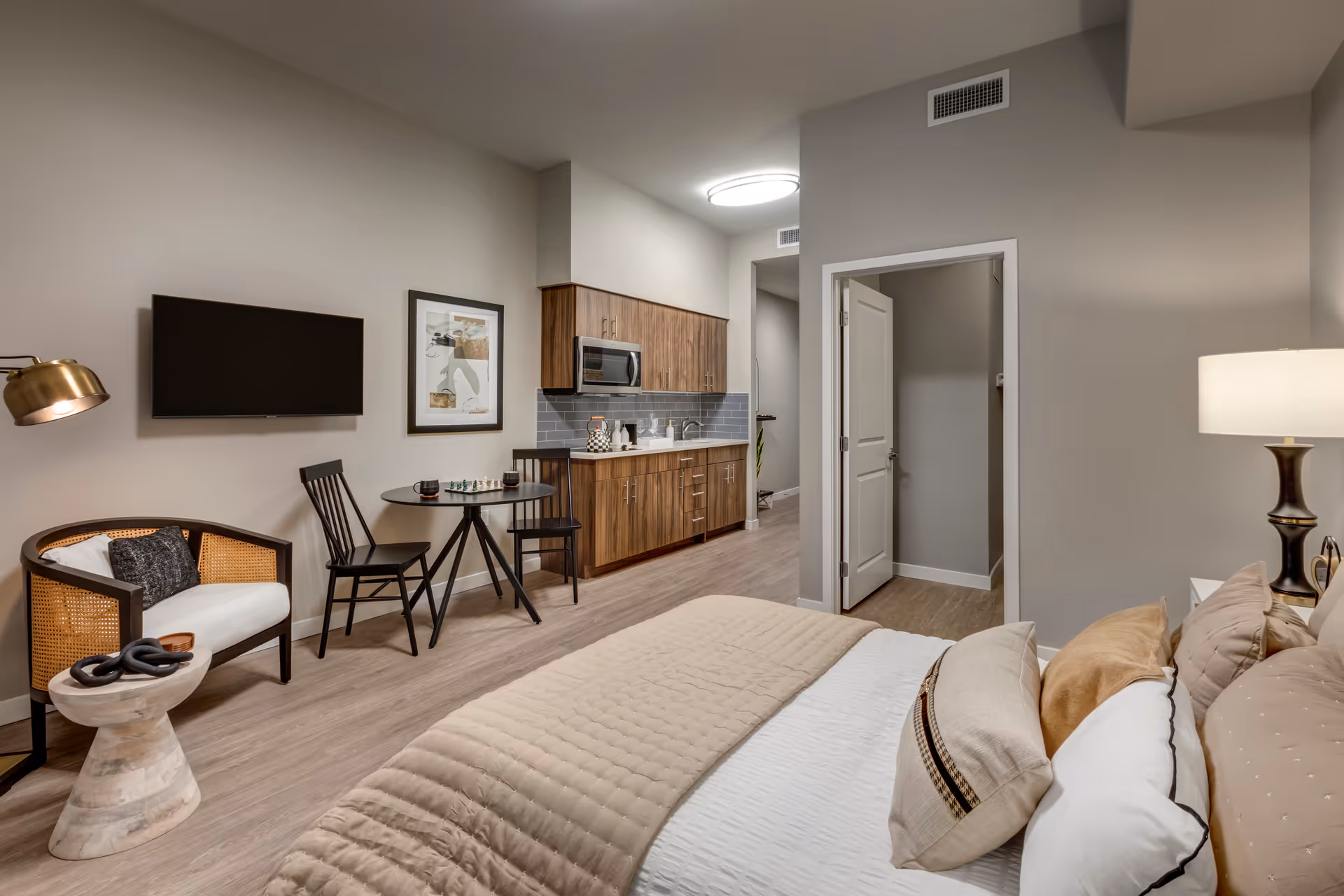Well-lit studio bedroom with a bed, bedside lamp, wall-mounted TV, small dining table and chairs, and a kitchenette along the back wall.