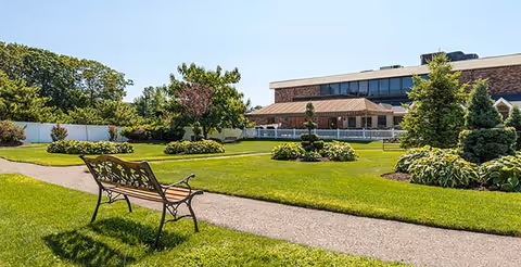 A well-maintained outdoor garden area with a paved walking path, a decorative metal bench, green grass, shrubs, and trees. In the background, there is a two-story building with large windows and a covered patio area.