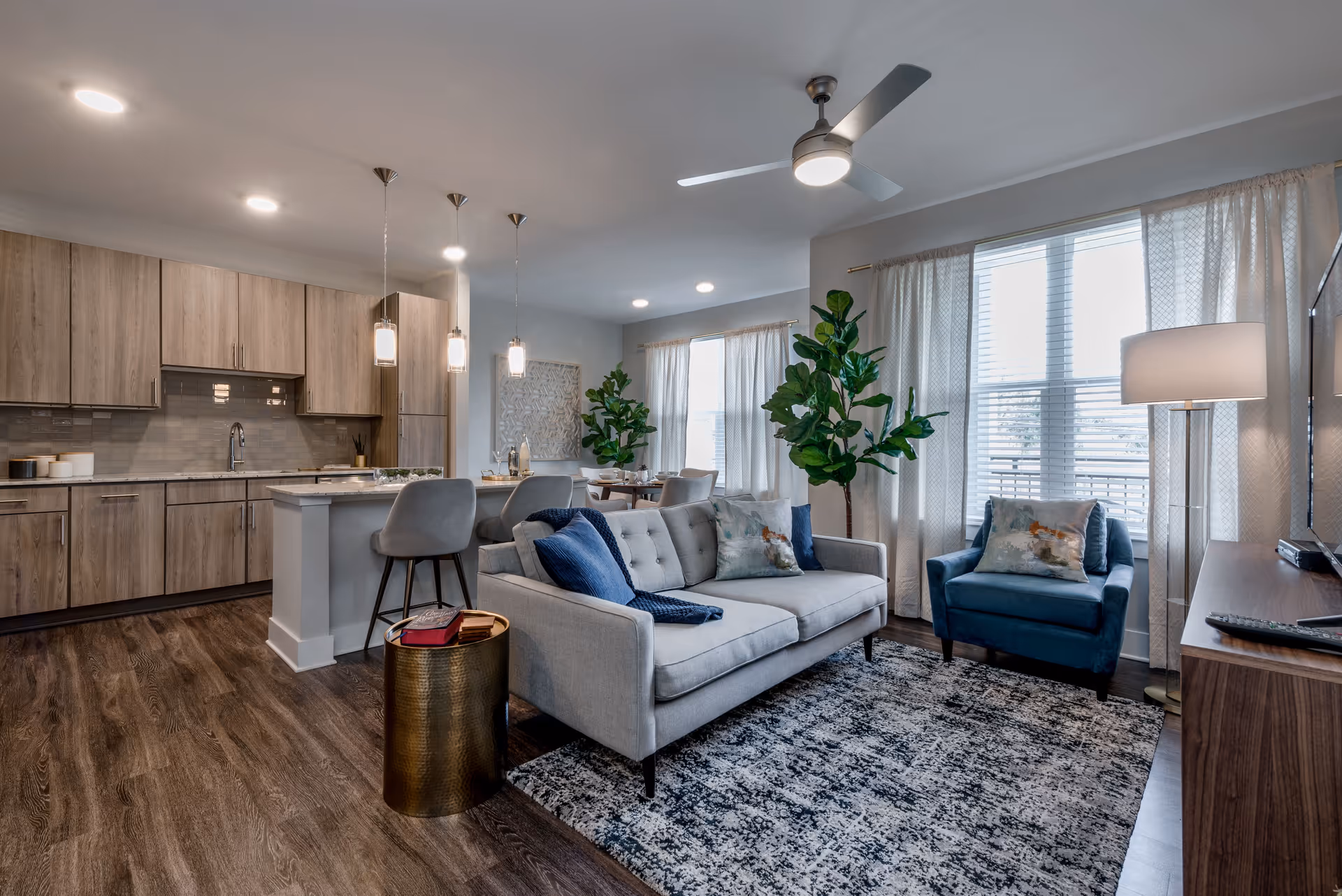 A modern open-concept living area featuring a light gray sofa with blue and patterned pillows, a blue armchair, a patterned rug, and a wooden TV stand with a flat-screen TV. The space includes a kitchen with light wood cabinets, a white island with two gray bar stools, and pendant lights hanging above. Large windows with white curtains allow natural light to fill the room, and there are two tall green plants adding a touch of nature.