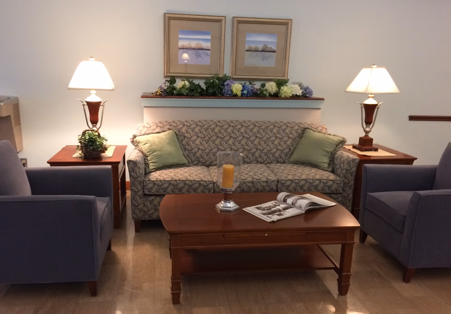 A furnished sitting area with a patterned sofa flanked by two armchairs, a wooden coffee table, side tables with lamps, and framed artwork above.