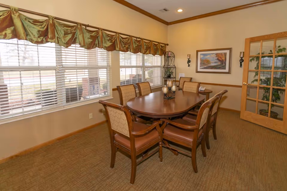 A dining room with a wooden oval table surrounded by six cushioned chairs. The room has large windows with blinds and a decorative valance, beige walls, a framed landscape painting, a small side table with a phone, a metal corner shelf with plants, and a wooden door with glass panels.