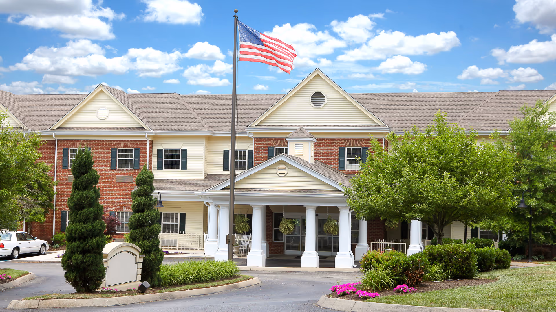 Exterior view of a senior living facility with a brick and beige siding facade, a covered entrance supported by white columns, an American flag on a flagpole in front, and well-maintained landscaping including trees, bushes, and flowers under a partly cloudy sky.