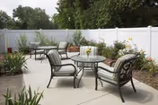 Outdoor patio area with glass-top tables, cushioned metal chairs, and plantings beside a white privacy fence.