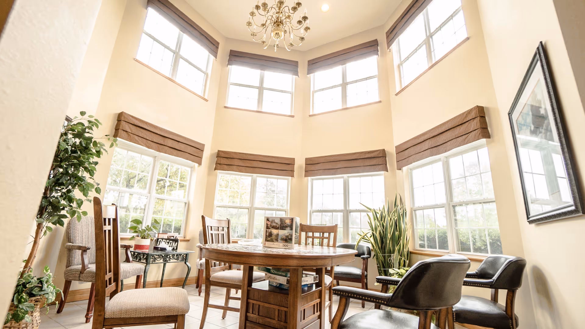 Sunlit common dining area with a round wooden table, chairs, tall bay windows, plants and a chandelier.