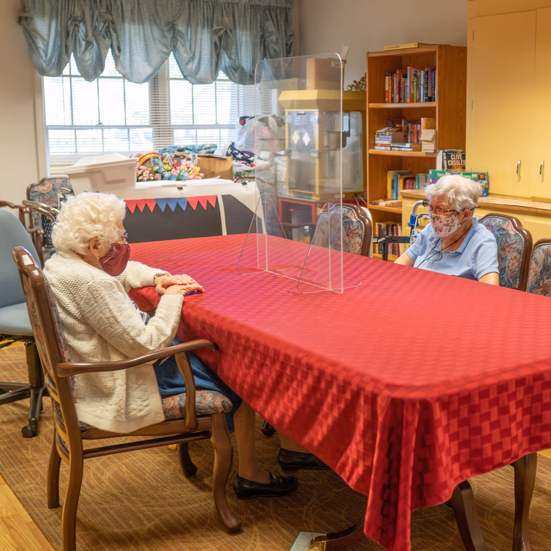Two elderly women wearing face masks sitting at a long table with a red tablecloth in a room with bookshelves and windows. A clear plastic divider is placed on the table between them.