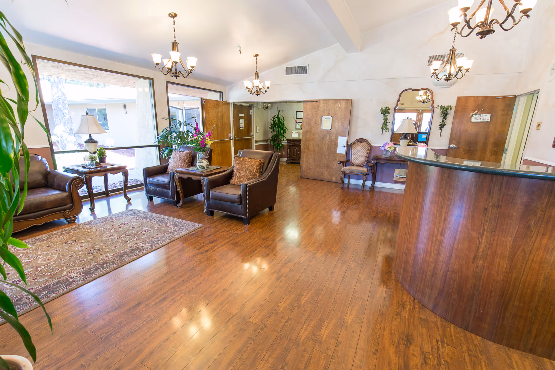 A spacious and well-lit reception area with wooden flooring, a curved wooden reception desk, leather armchairs, a side table with a lamp, a decorative rug, plants, and chandeliers hanging from the ceiling. There are large windows letting in natural light and wooden doors leading to other rooms.