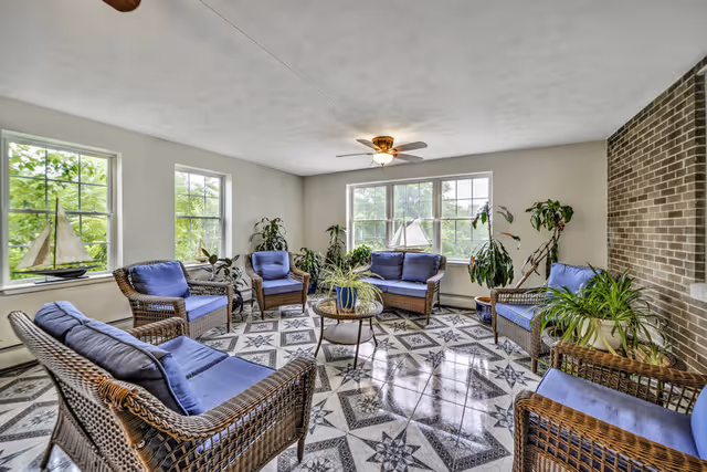 A bright living room with large windows showing greenery outside. The room features wicker furniture with blue cushions arranged in a circle around a glass-top coffee table. Several potted plants are placed around the room, and a ceiling fan with a light is mounted on the white ceiling. The floor has a patterned tile design, and a brick accent wall is visible on the right side.
