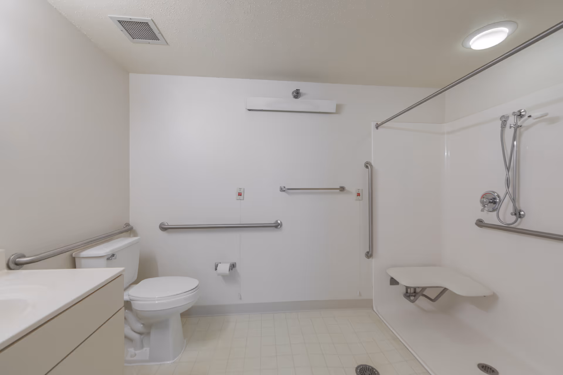 A clean, accessible bathroom with white walls and flooring. It features a toilet with grab bars on the wall behind and beside it, a sink with a countertop on the left, and a walk-in shower area on the right with a fold-down seat, grab bars, and a handheld showerhead. The ceiling has a vent and a round light fixture.
