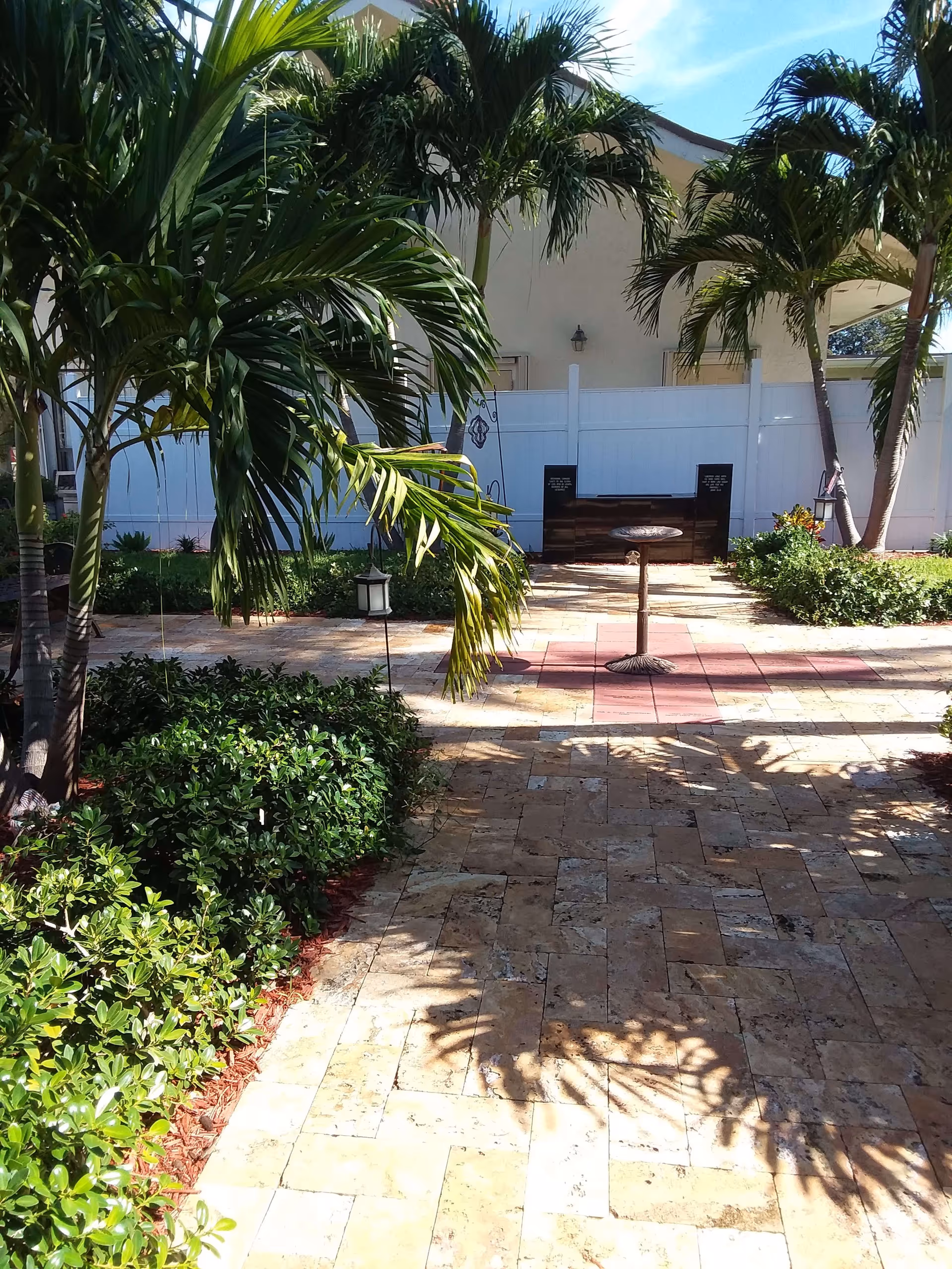 A sunny outdoor garden area with palm trees, green shrubs, and a paved stone walkway leading to a small birdbath or decorative stand. There is a white fence and a beige building in the background.