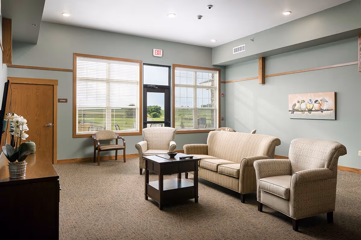 A cozy senior living facility common area with a beige sofa, two matching armchairs, a wooden coffee table, and a wooden chair near large windows. The walls are painted light green with wooden trim, and there is a painting of birds on a branch hanging on the wall. A door with an exit sign above it leads outside, and a wooden cabinet with a potted orchid is visible on the left.
