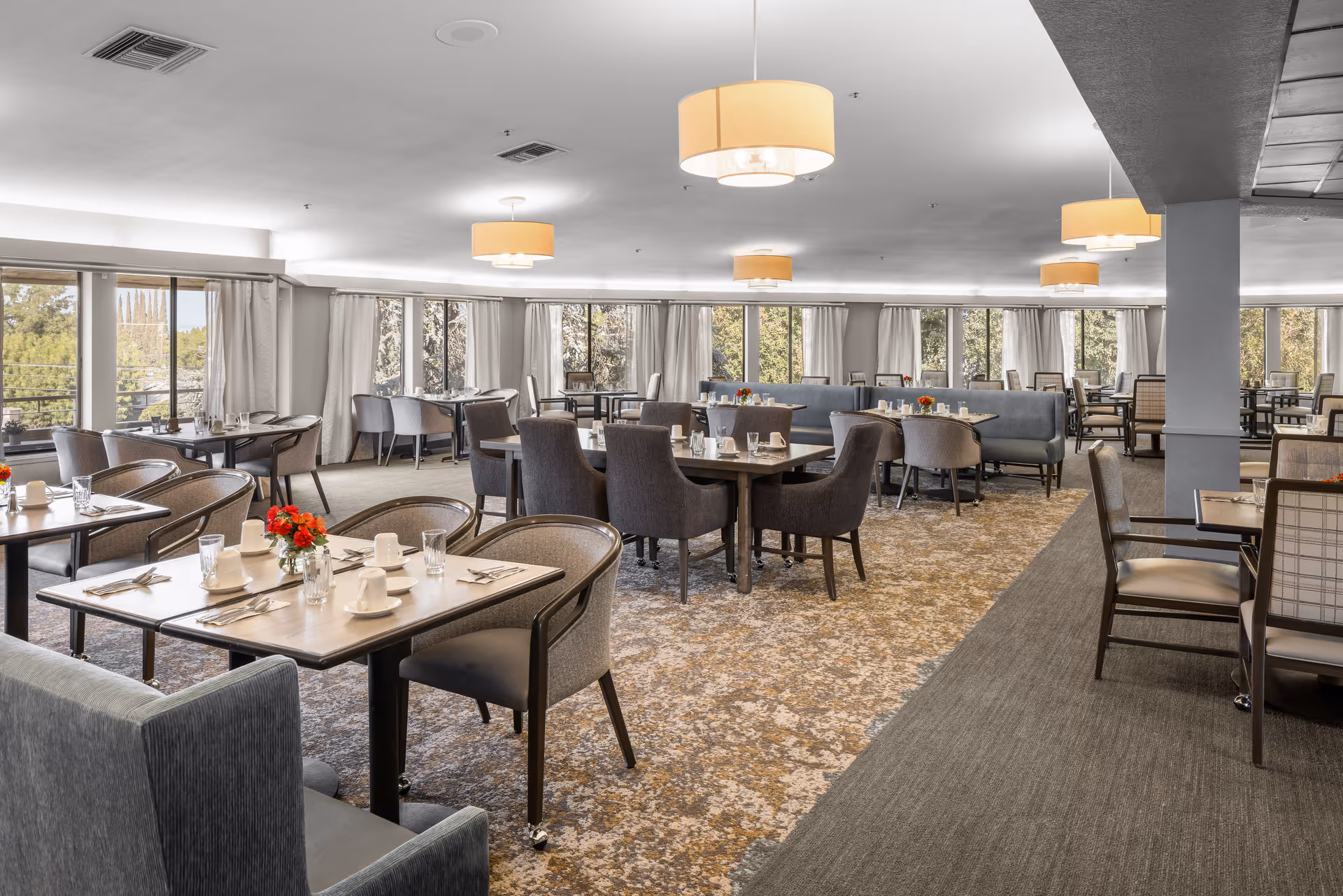 A spacious dining room in a senior living community with multiple tables set with cups, glasses, and silverware. The room features large windows with curtains allowing natural light to fill the space, modern pendant lights hanging from the ceiling, and a mix of upholstered chairs and booth seating. The carpet has a patterned design, and small flower arrangements are placed on the tables.