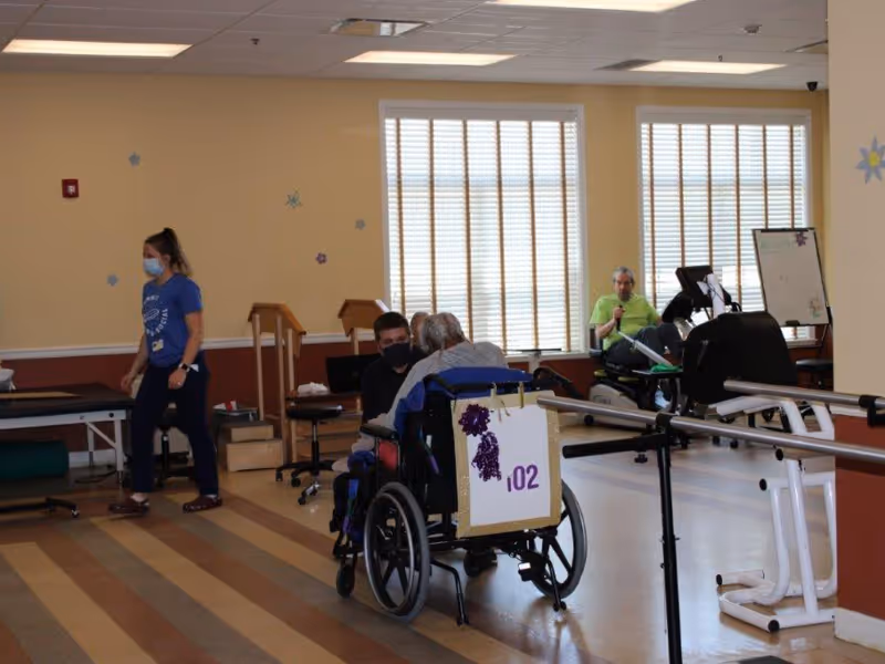 A rehabilitation room with two elderly individuals using exercise equipment and a wheelchair. A staff member wearing a mask and blue shirt is walking nearby. The room has large windows with blinds, exercise machines, and parallel bars for physical therapy.