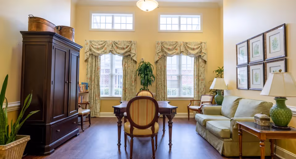 Bright, traditionally furnished living room with a sofa, wooden armoire, table and chairs in front of large windows and decorative lamps.