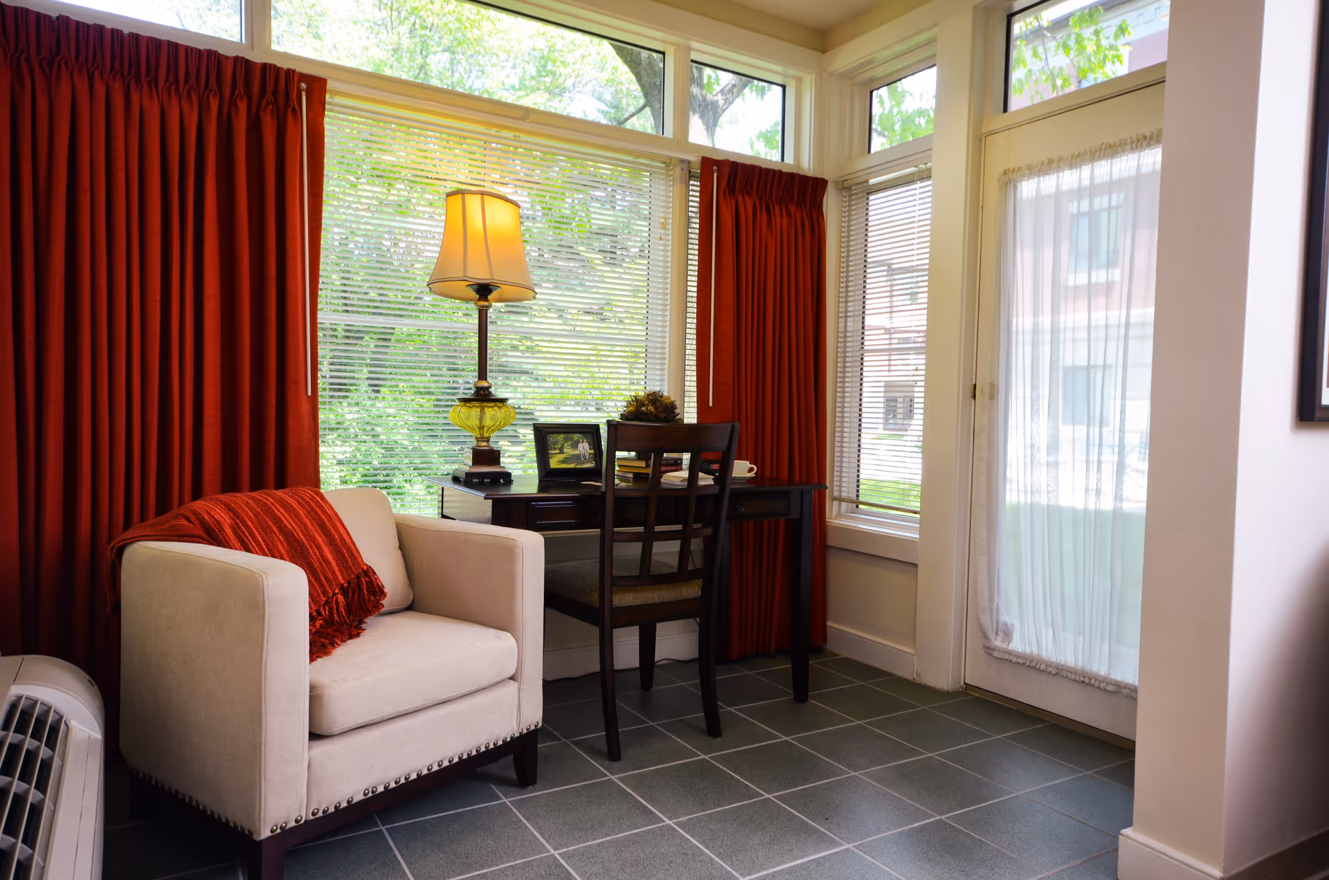 A cozy corner of a senior living community room featuring a beige armchair with a red throw blanket, a dark wooden desk with a chair, a table lamp, and framed pictures. Large windows with red curtains and blinds allow natural light to fill the space, and a glass door with a sheer curtain leads outside.