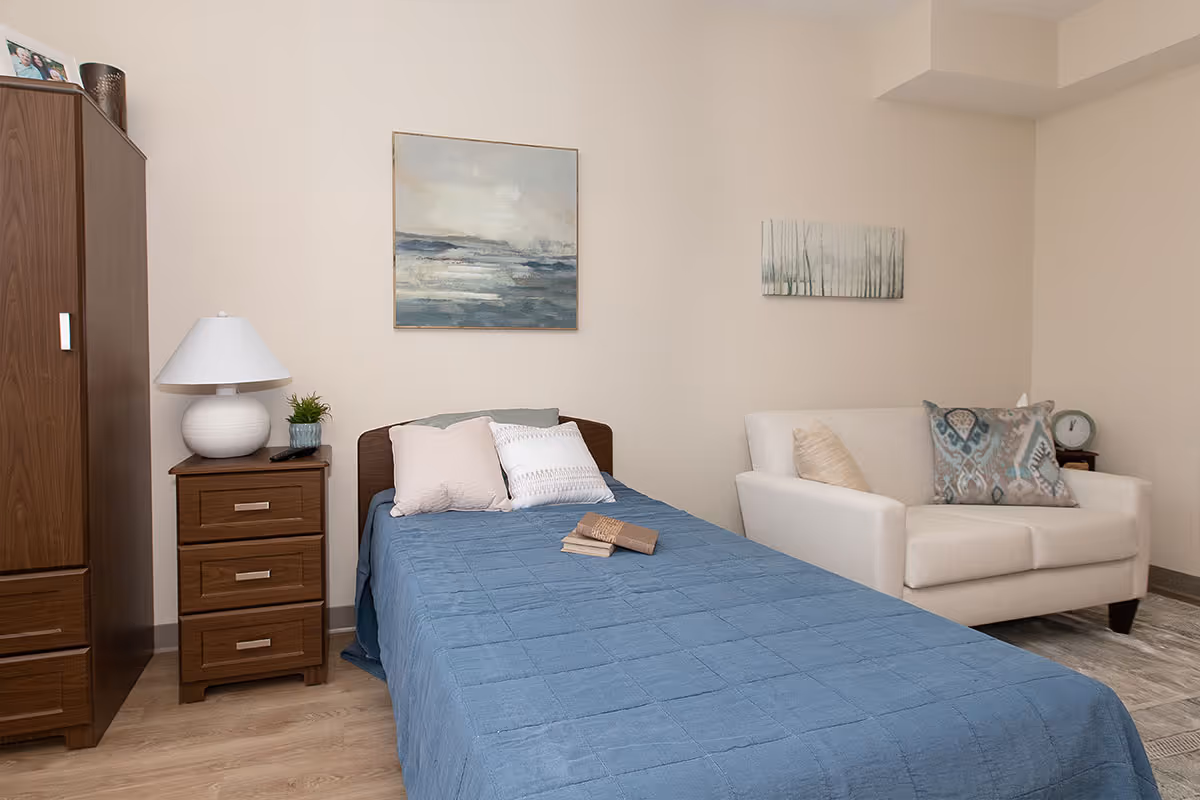 Furnished bedroom with a single bed covered by a blue quilt, a wooden nightstand and wardrobe, wall art, and a small sofa.