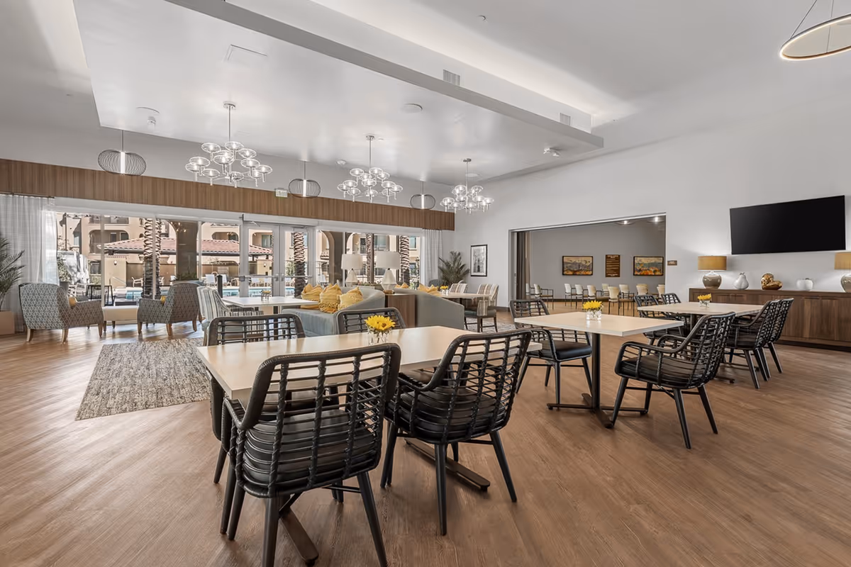 A spacious and modern common area in a senior living facility with multiple tables and black chairs arranged neatly. The room features large windows with a view of an outdoor pool area and palm trees. There are comfortable armchairs, a sofa with yellow pillows, contemporary chandeliers hanging from the ceiling, a large flat-screen TV mounted on the wall, and decorative lamps and vases on a wooden cabinet.