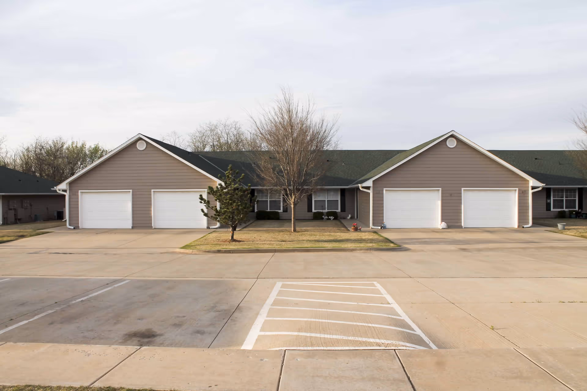 Exterior view of a single-story residential building with multiple garages and a central entrance, surrounded by a paved parking area and small trees on the lawn.