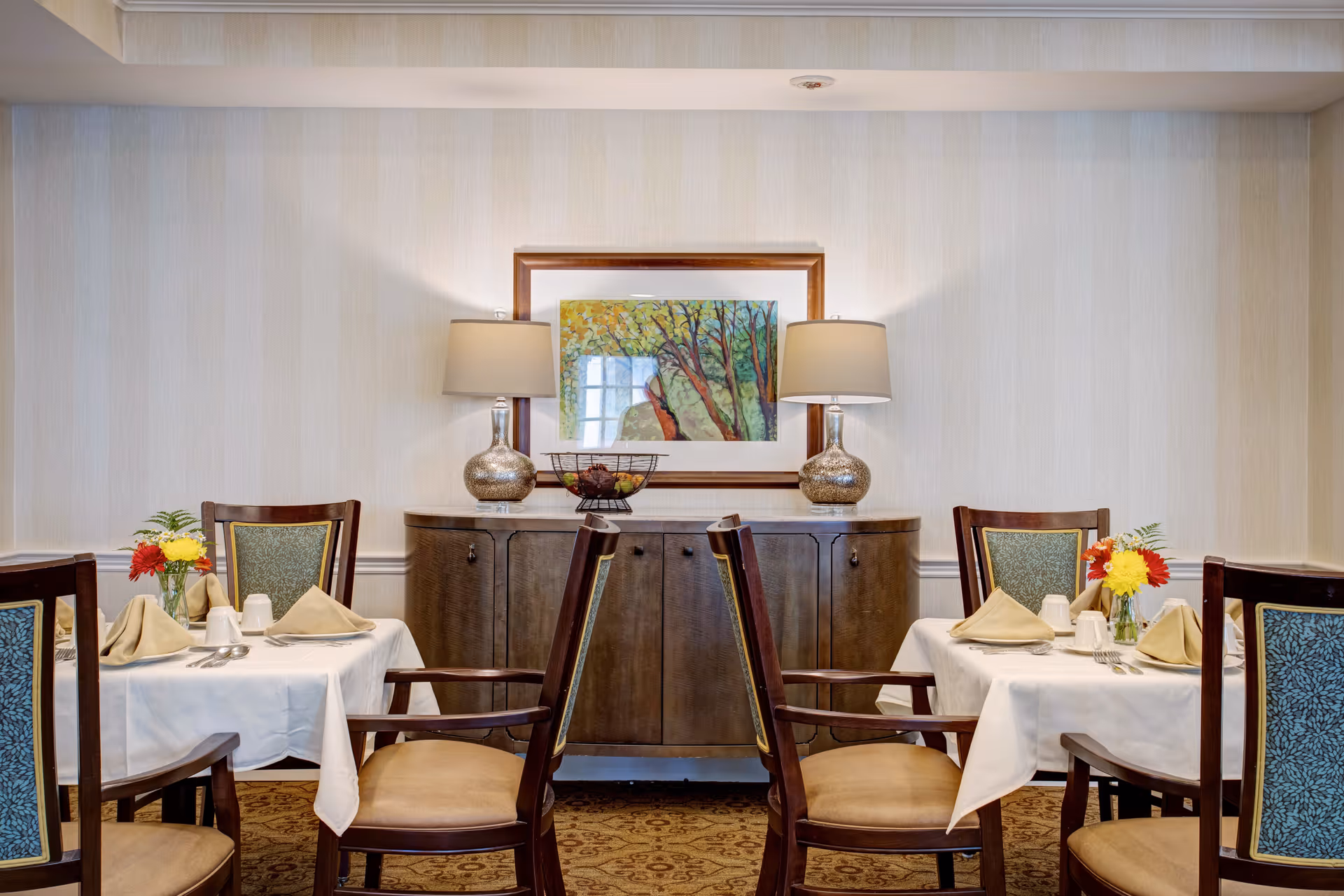 A dining room with two tables set for four people each, featuring white tablecloths, beige napkins, silverware, and small flower arrangements. Behind the tables is a wooden sideboard with two decorative lamps and a framed painting of trees on the wall above it.