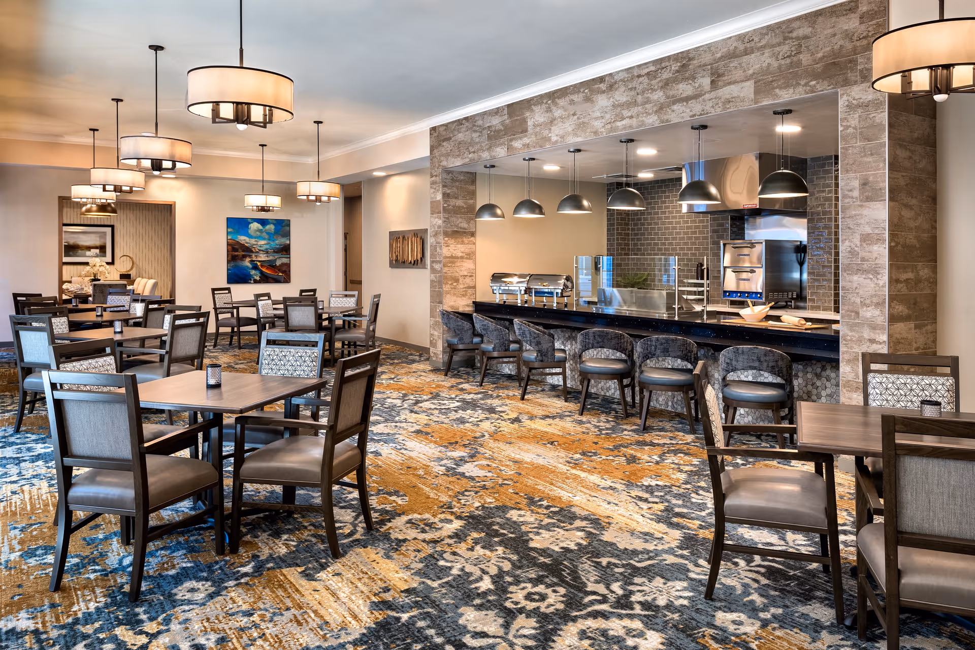 A spacious dining room in a senior living facility featuring multiple square wooden tables with cushioned chairs arranged neatly. The room has a patterned carpet in shades of blue, gold, and beige. On the right side, there is a serving counter with bar stools and pendant lights hanging above. The back wall has a tiled backsplash and stainless steel kitchen equipment visible through the serving window. The ceiling has several modern drum pendant lights, and there are paintings and decorative wall art on the walls.