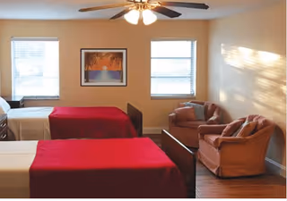 A senior living bedroom with two single beds covered in red blankets, a ceiling fan with lights, two windows with blinds, a framed picture on the wall, and two cushioned armchairs in the corner.