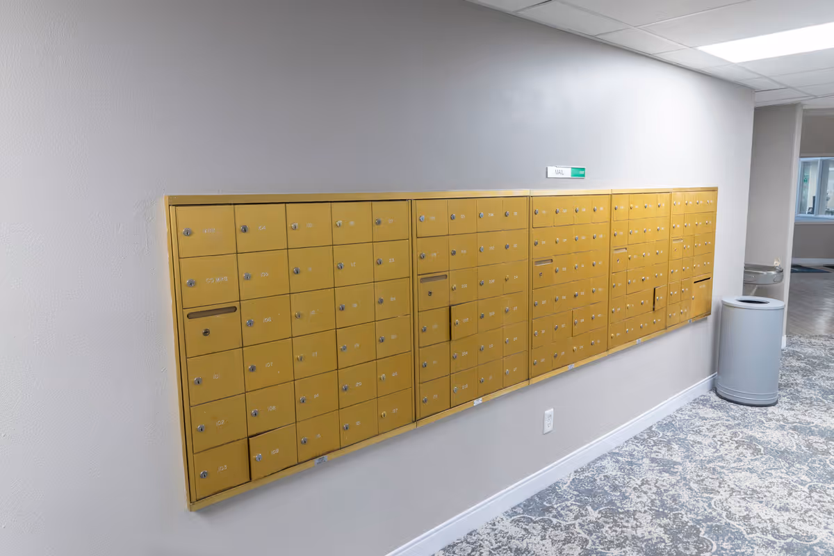 A wall-mounted set of yellow mailboxes with multiple locked compartments, each labeled with numbers. The mailboxes are installed on a light gray wall above a patterned carpeted floor. A trash can is positioned near the mailboxes, and a sign above the mailboxes reads 'MAIL OUT'.