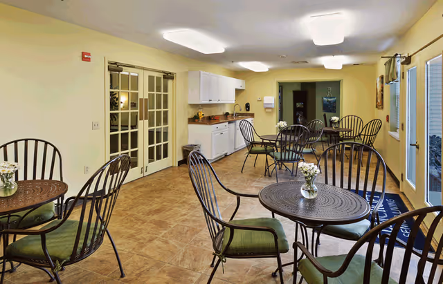 Communal dining room with round metal tables and green-cushioned chairs, a small kitchenette, and French doors.