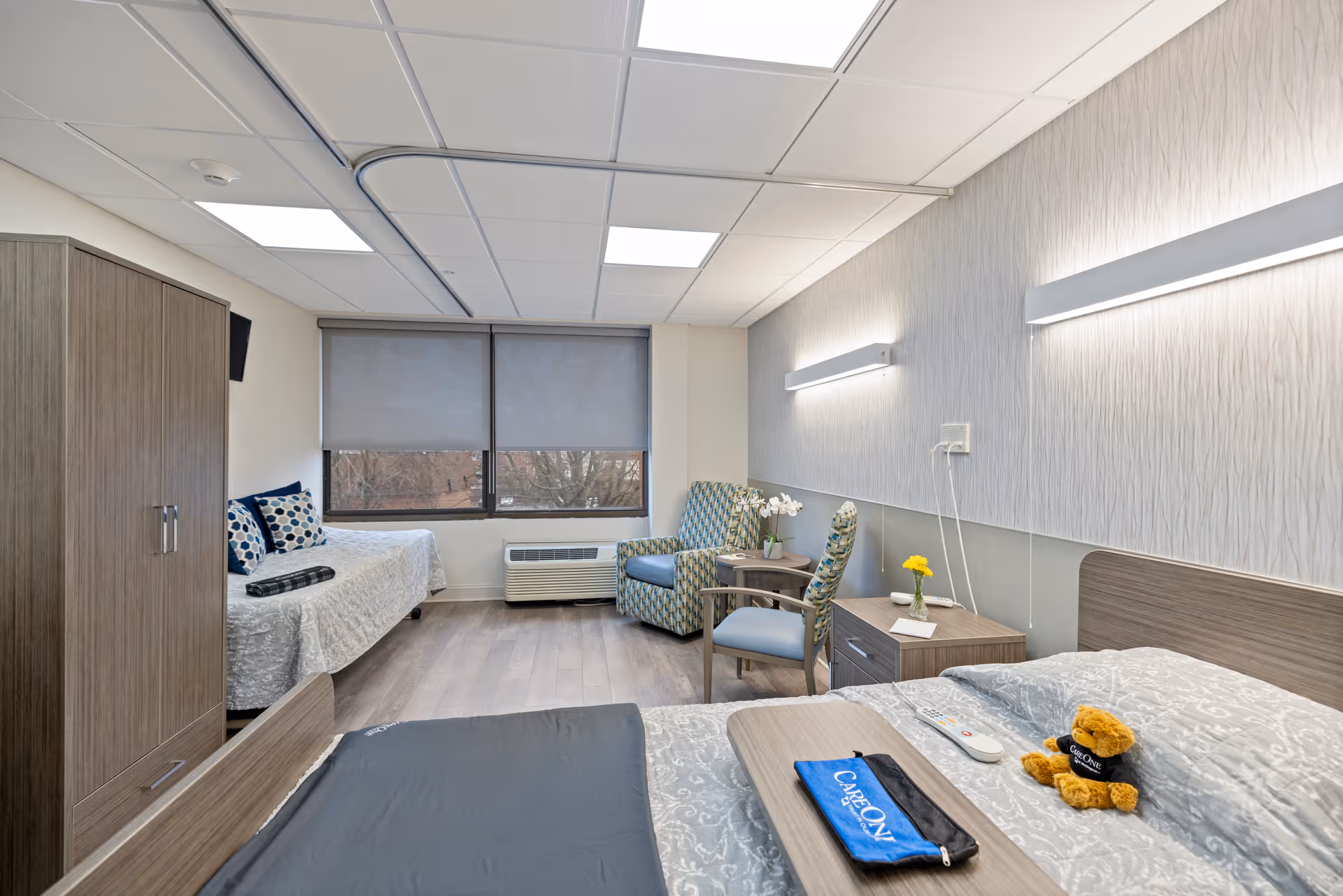 A bright and clean senior living bedroom with two beds, one near the window and one in the foreground. The room features wood flooring, a wooden wardrobe, two patterned armchairs with a small round table between them, and a large window with closed blinds. On the bed in the foreground, there is a small teddy bear wearing a CareOne shirt, a remote control, and a blue pouch with the CareOne logo. The walls have modern light fixtures and a textured wallpaper.