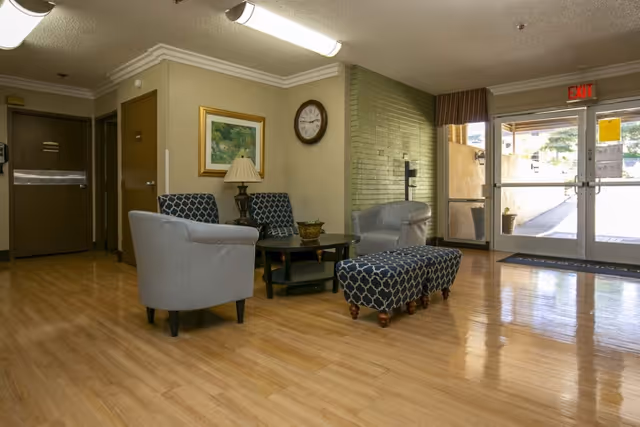 A seating area in a facility lobby with two gray armchairs, two blue patterned chairs, a matching blue patterned ottoman, a black coffee table, a table lamp, a wall clock, and a framed painting. The floor is wooden, and there is a glass exit door leading outside.
