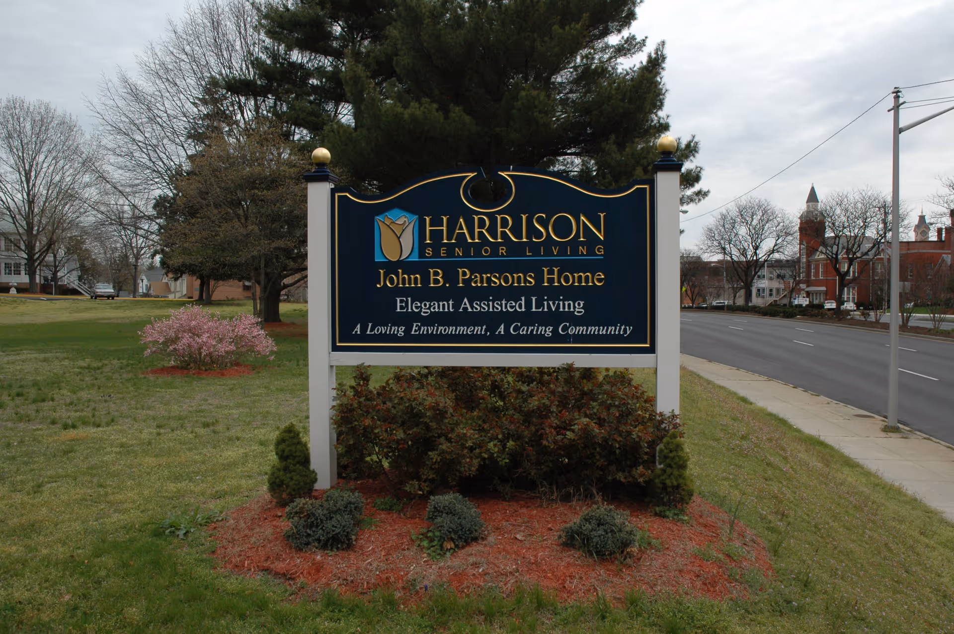 Outdoor view of a sign for Harrison Senior Living, John B. Parsons Home, Elegant Assisted Living, located on a grassy area next to a road with trees and buildings in the background.