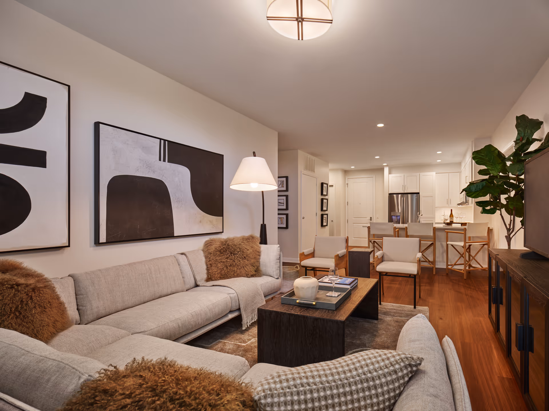A modern living room with a light gray sectional sofa adorned with brown and patterned pillows, a dark wooden coffee table with decorative items, two beige armchairs, and a floor lamp. The room features abstract black and white artwork on the wall, hardwood flooring, and an open view into a kitchen area with white cabinetry, a stainless steel refrigerator, and a breakfast bar with wooden stools. A large green plant is placed near a TV on a dark wooden stand.