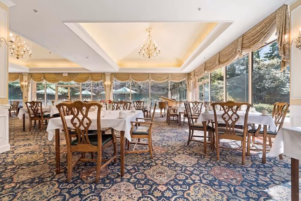 Bright, formal dining room with tables set in white linens, wooden chairs, chandeliers, and large windows overlooking greenery.