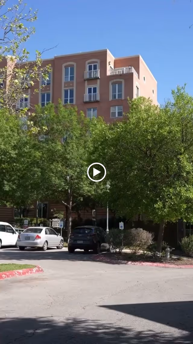 Exterior view of a multi-story brick building partially obscured by green trees. Several cars are parked in front of the building under a clear blue sky.