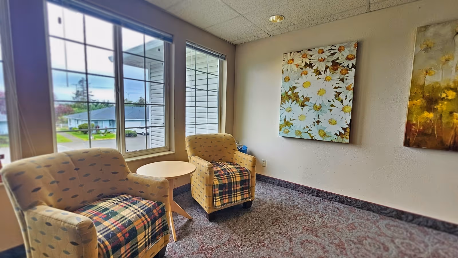 Two upholstered armchairs with a small round table by large windows and floral wall art in a sitting area.